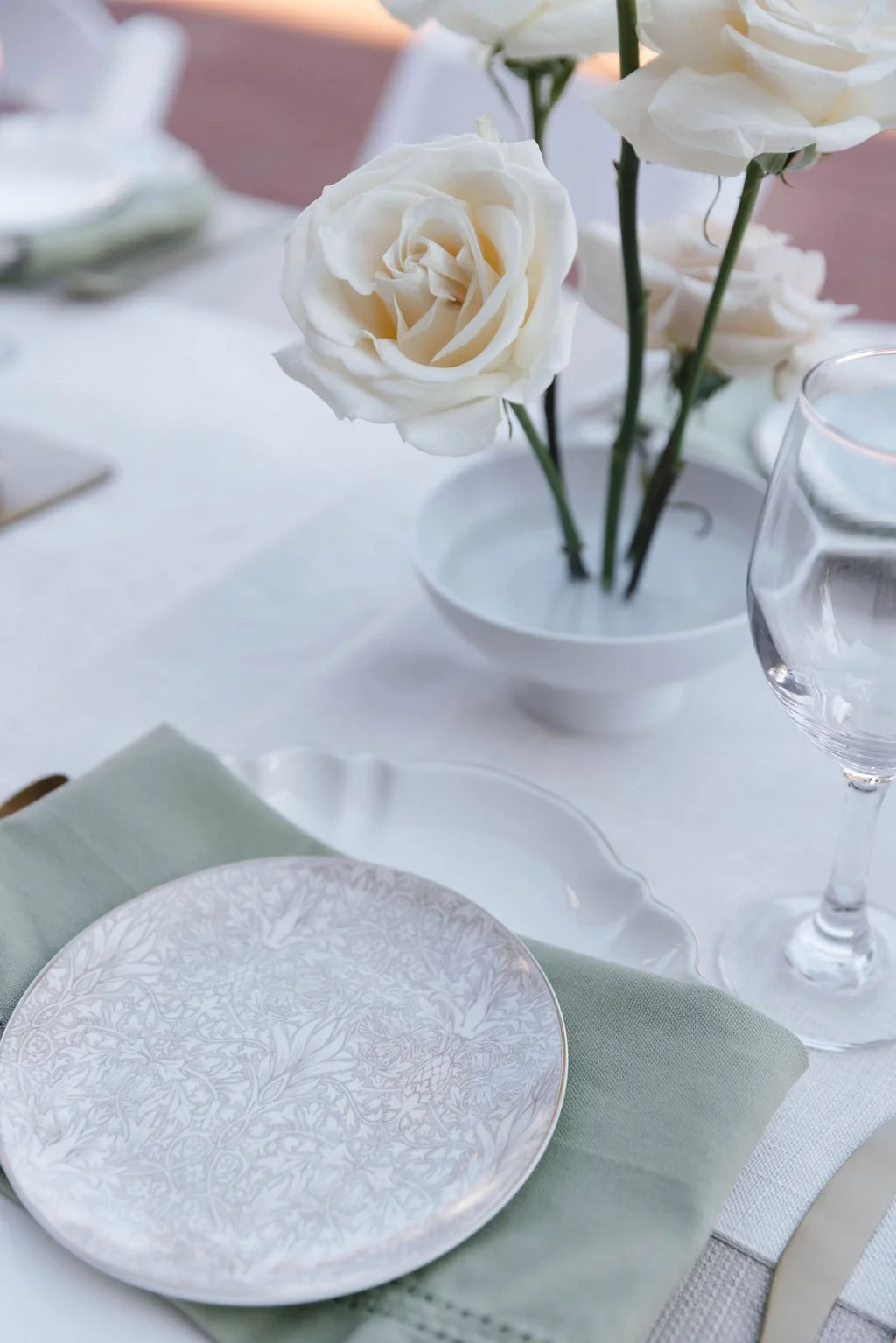 Elegant table setting with a white rose floral centerpiece in a white vase, a decorative patterned plate on a light green napkin, a wine glass, and a water glass, all arranged on a white tablecloth.