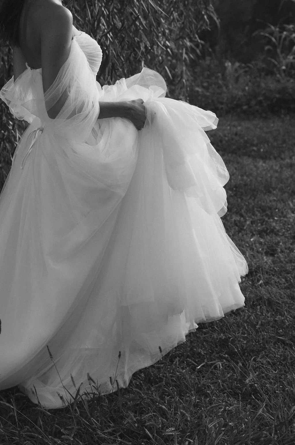 A woman in a flowing, strapless wedding gown standing outdoors on grass, with tall plants in the background. The image is in black and white.