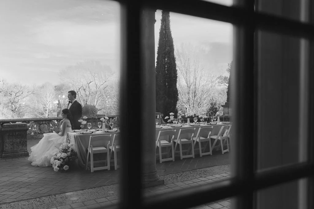A black and white photo of a wedding reception outdoors, with a bride and groom near a long table set with glasses and flowers, seen through a window.