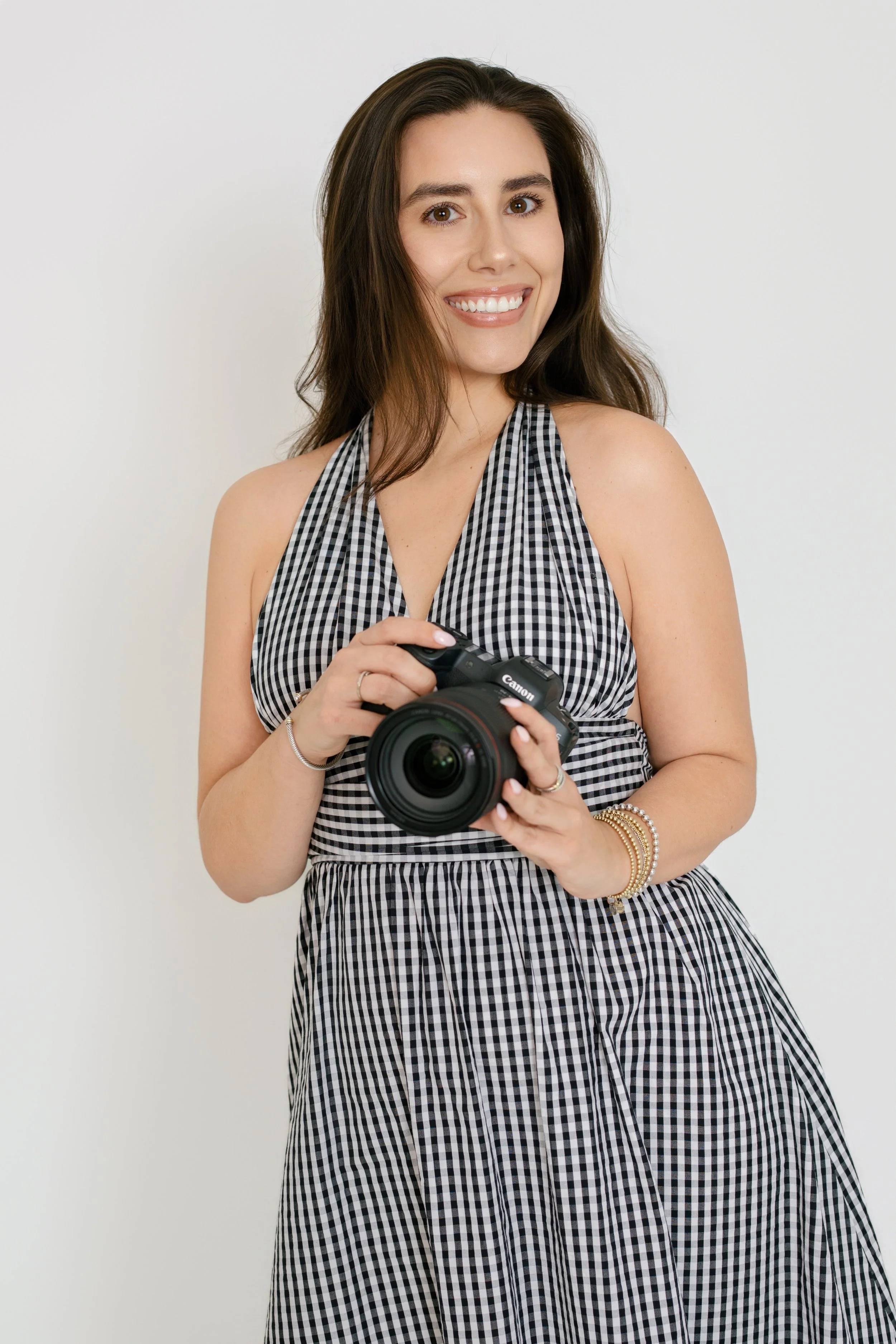 A woman with long brown hair, smiling, holding a Canon camera, wearing a black and white gingham dress with accessories, standing against a plain white background.