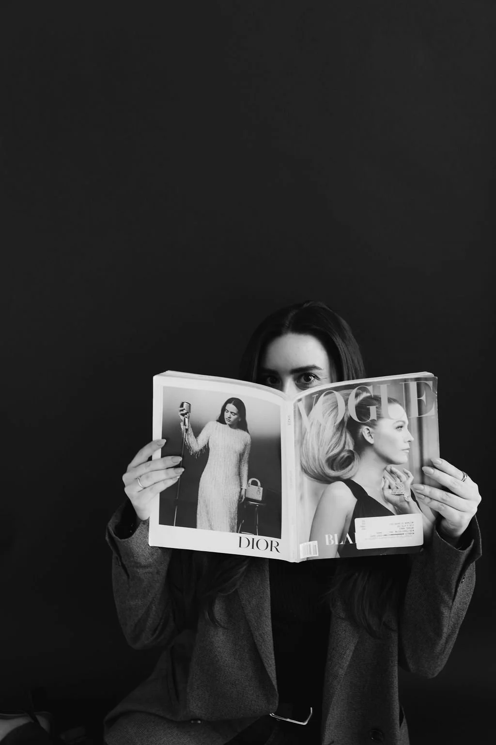 A woman holding a Vogue magazine with a black and white fashion photograph on the cover, partially obscuring her face, against a dark background.