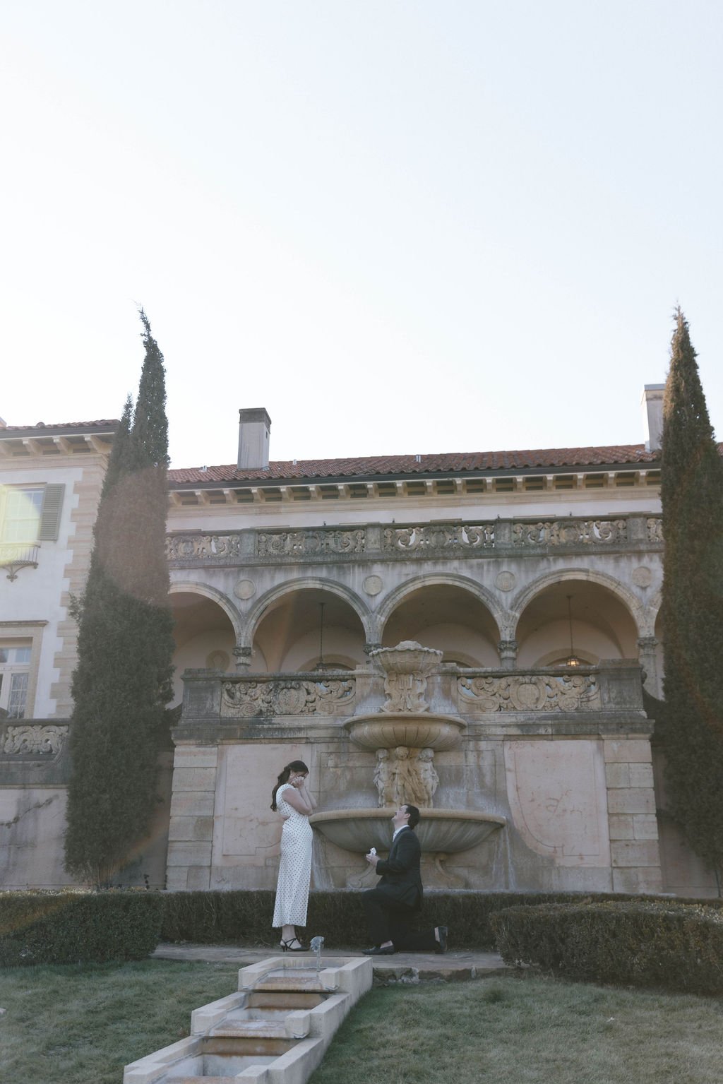 A man proposing to a woman in front of a decorative stone fountain on a well-maintained lawn, with tall cypress trees and an elegant historic building in the background.