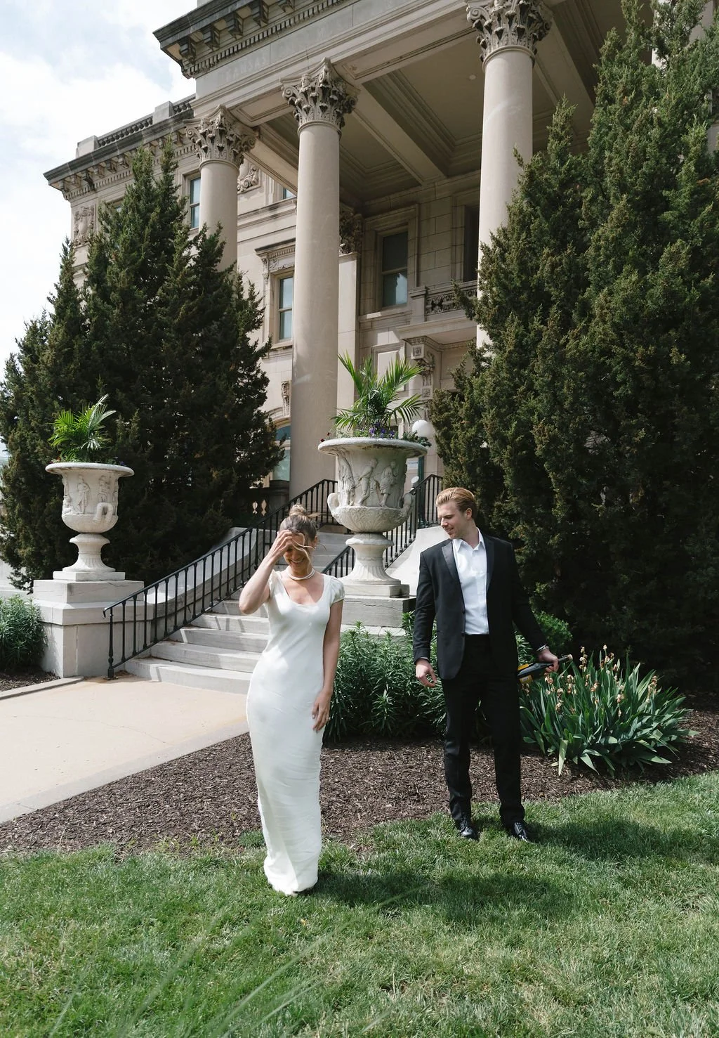 A woman in a white dress and a man in a black suit walking outside a large, ornate building with tall columns, surrounded by greenery and decorative planters.