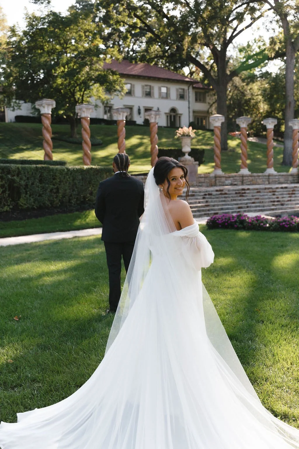 A bride in a white wedding gown and veil smiling at the camera, with a groom in a black suit walking away in the background, outdoors on a sunny day near a mansion and a lush, green lawn.