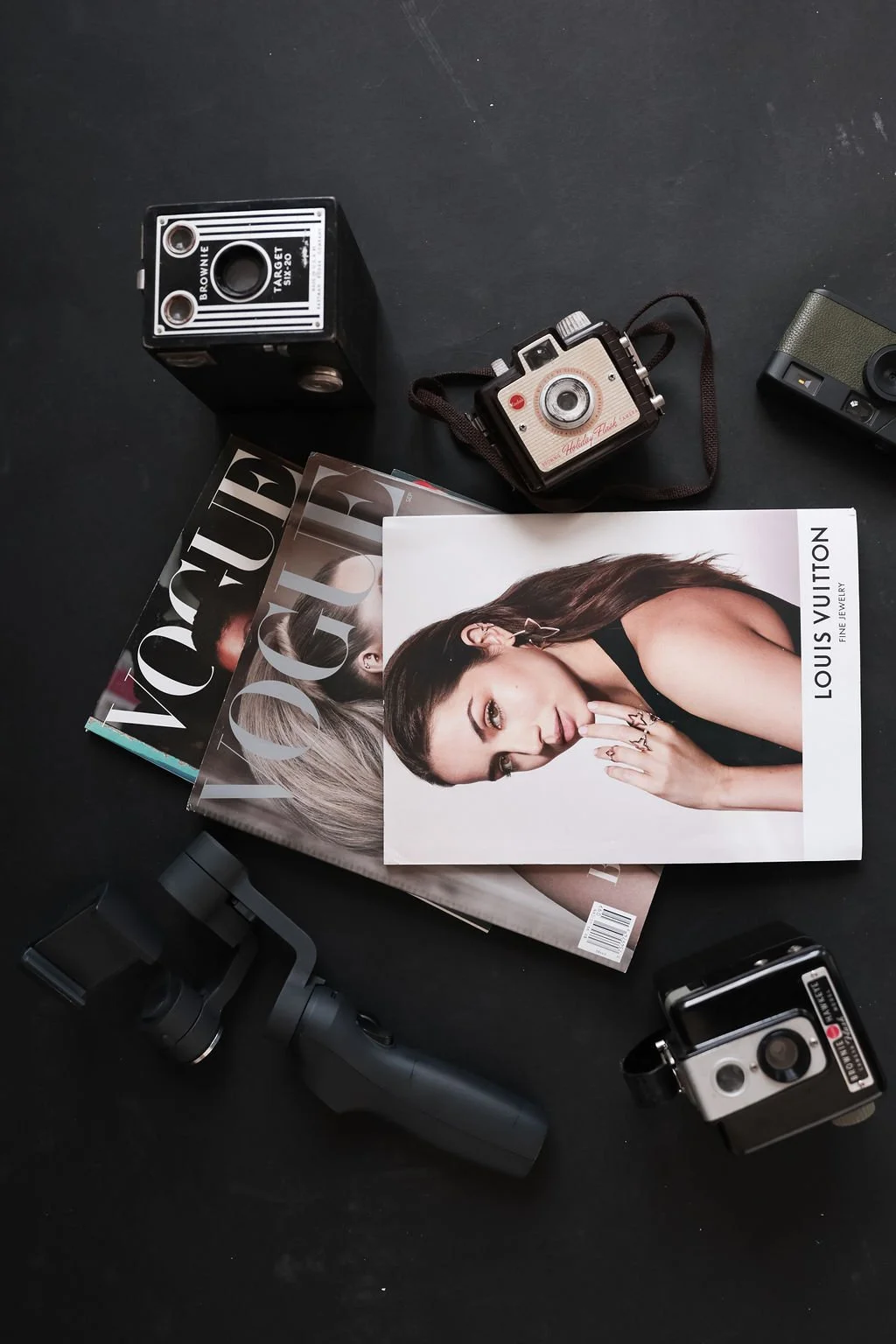 A flat lay of fashion magazines, vintage cameras, a stabilizer, and a camera phone on a black surface.