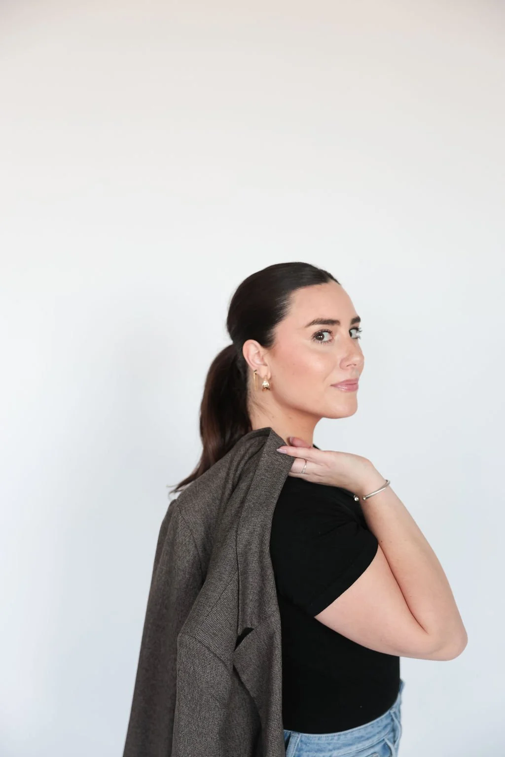 A woman with dark hair tied back, wearing a black shirt and blue jeans, is holding a brown blazer over her shoulder with her left hand, standing against a plain white wall.