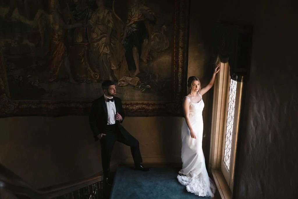 A bride in a white wedding gown and a groom in a tuxedo inside a dimly lit room with a large tapestry on the wall. The bride is standing near a window, touching the curtain, while the groom is standing a few feet away, looking at her.