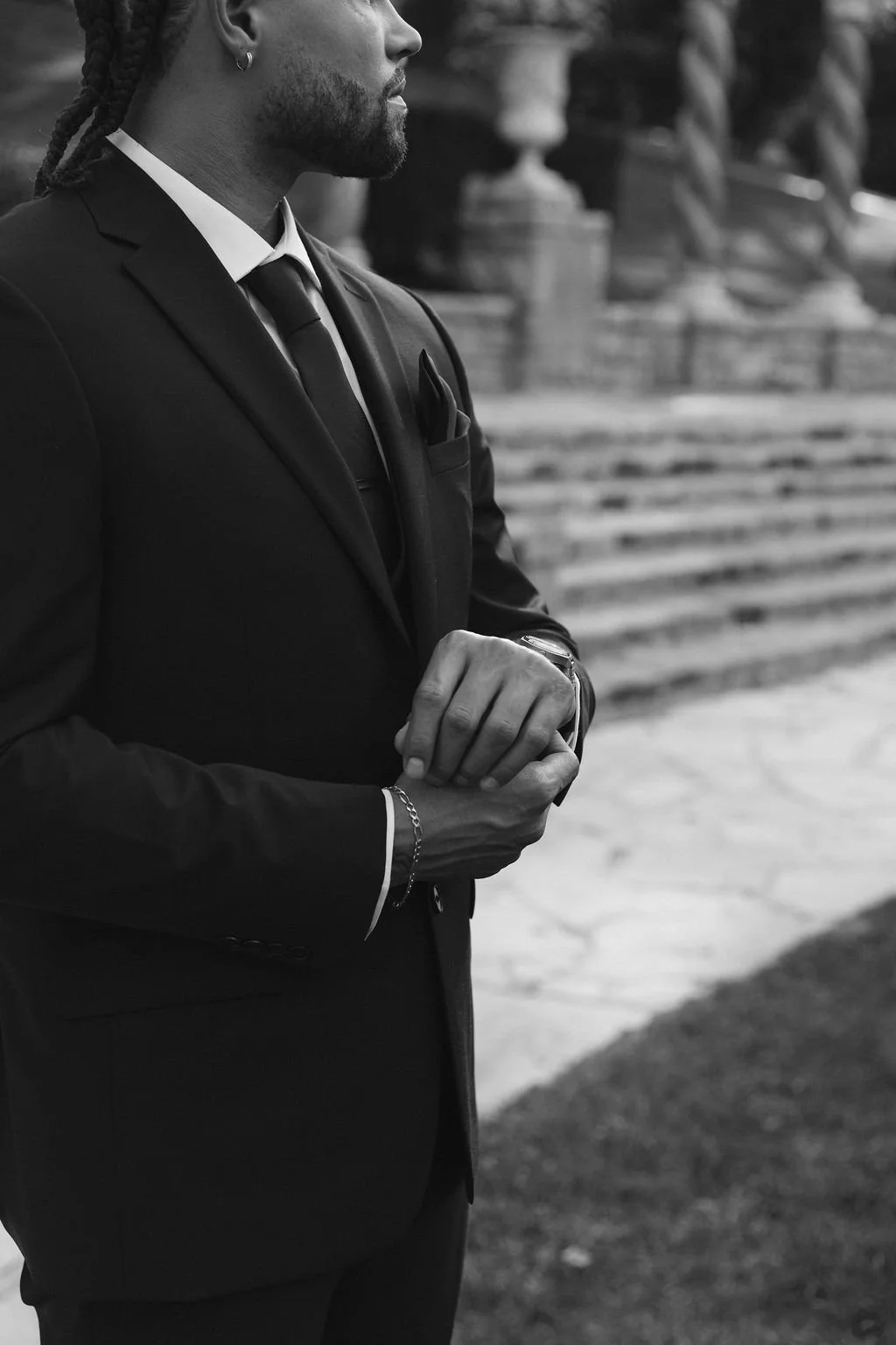 A man in a dark suit and tie stands outdoors with his hands clasped, wearing a watch and bracelet, with decorative stone steps and columns in the background.