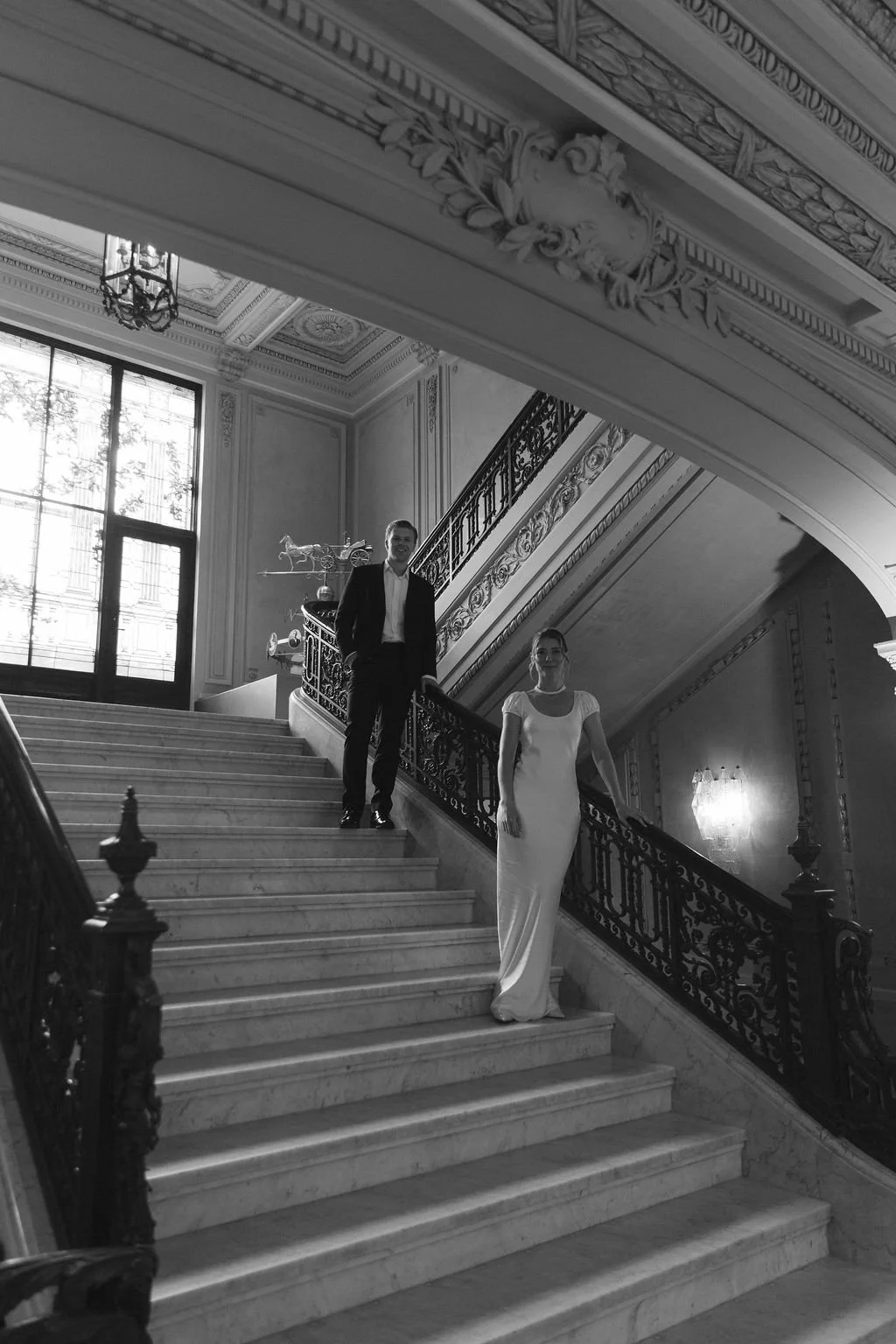 A man in a suit and a woman in a white gown standing on a grand marble staircase in an ornate, historic building with tall windows and decorative molding.