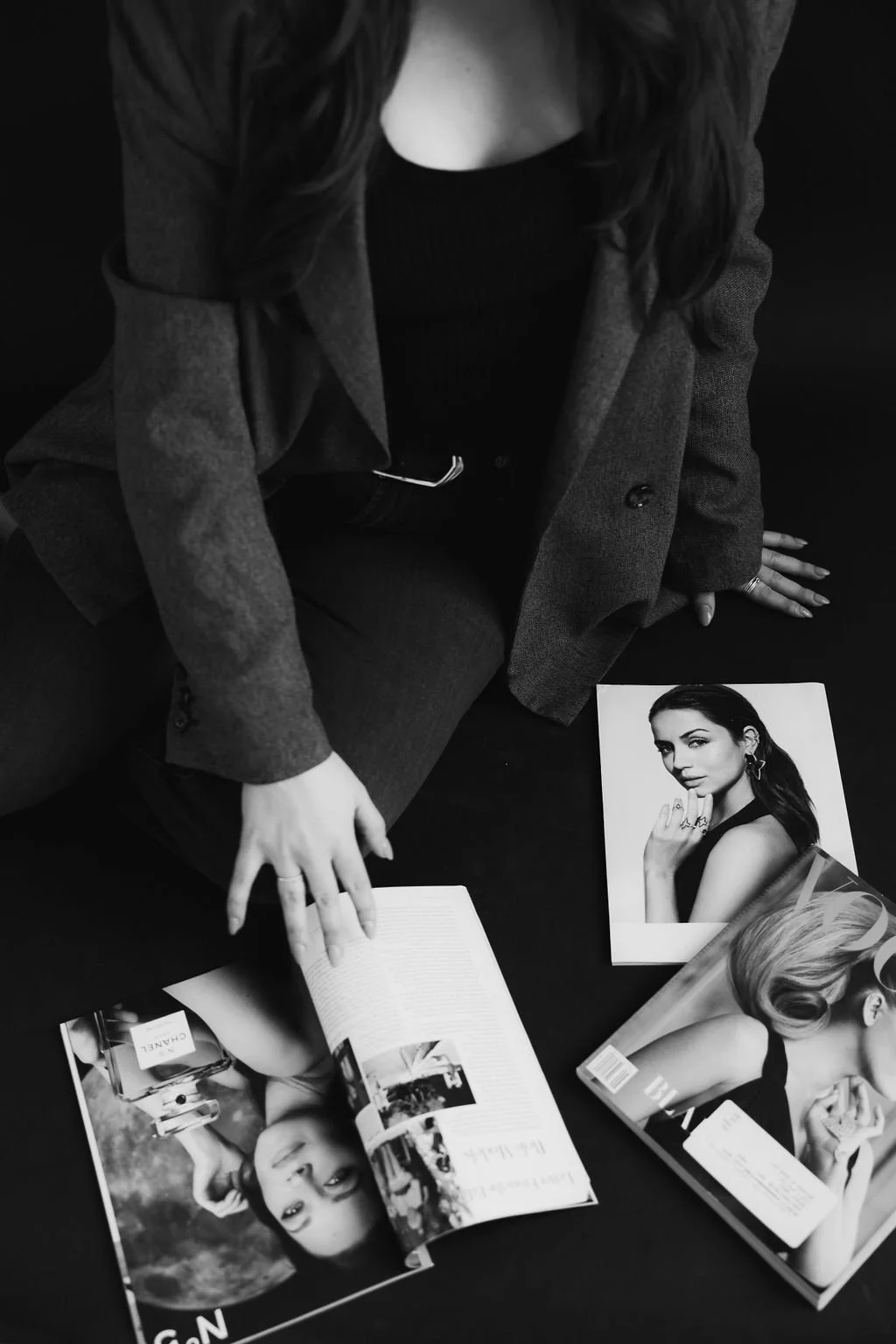 A woman with dark hair wearing a blazer sits on the floor, surrounded by fashion magazines, with a magazine open and photographs of models visible.