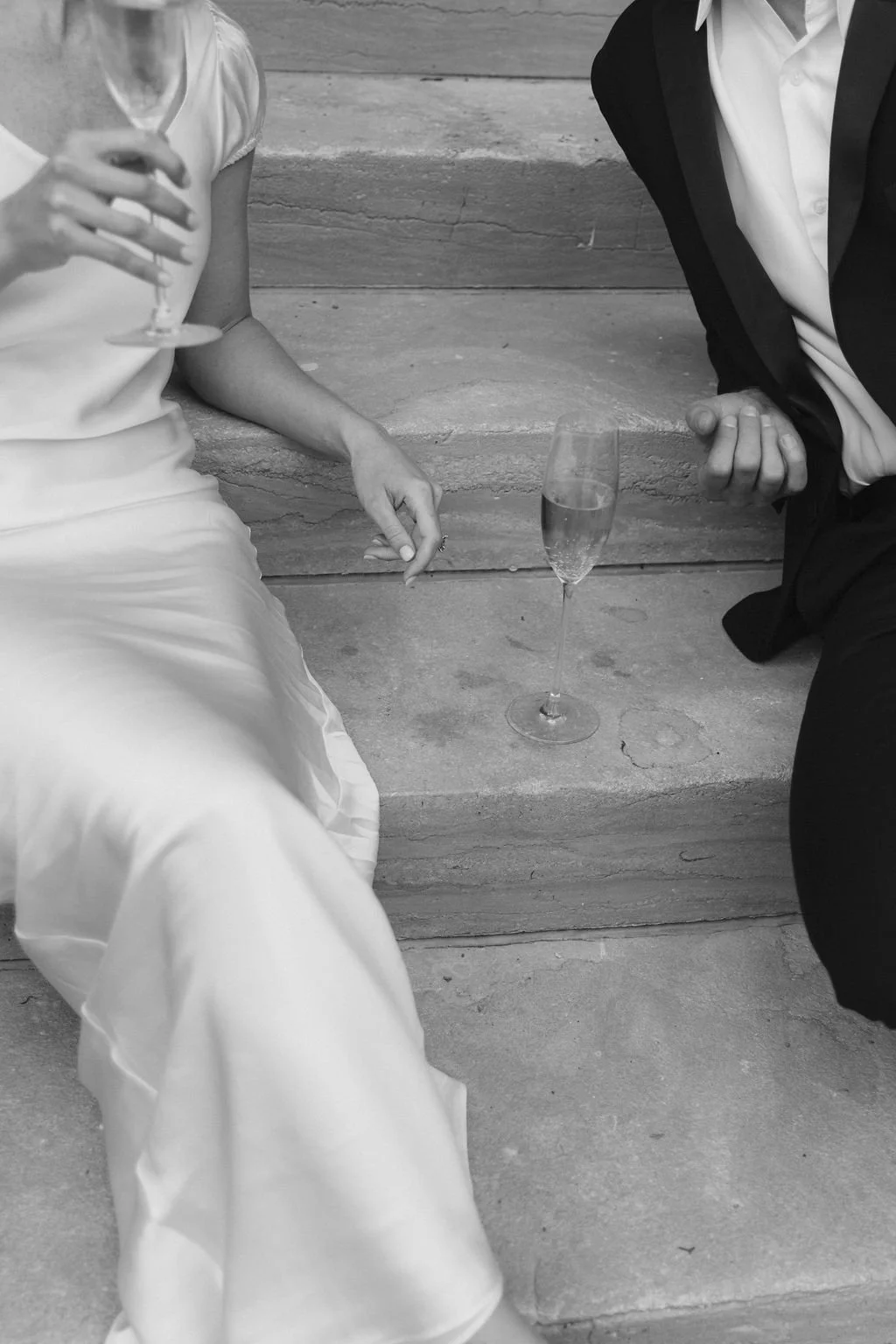 Close-up of two people celebrating with Champagne, sitting on stone steps. The woman is wearing a white dress and holding a glass of champagne, while the man is dressed in a suit with an open collar, and has a glass of champagne on the steps between them.