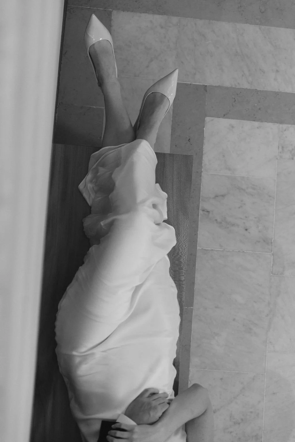 A person lying on the floor with legs crossed, wearing white pointed shoes and a long white dress, seen from above in black and white.