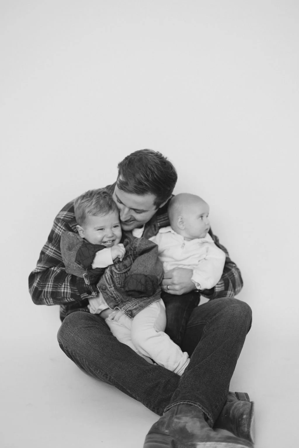 A man sitting on the floor holding two young children, one on each side, with a plain background. The man is smiling and interacting with the children, who appear happy and relaxed.