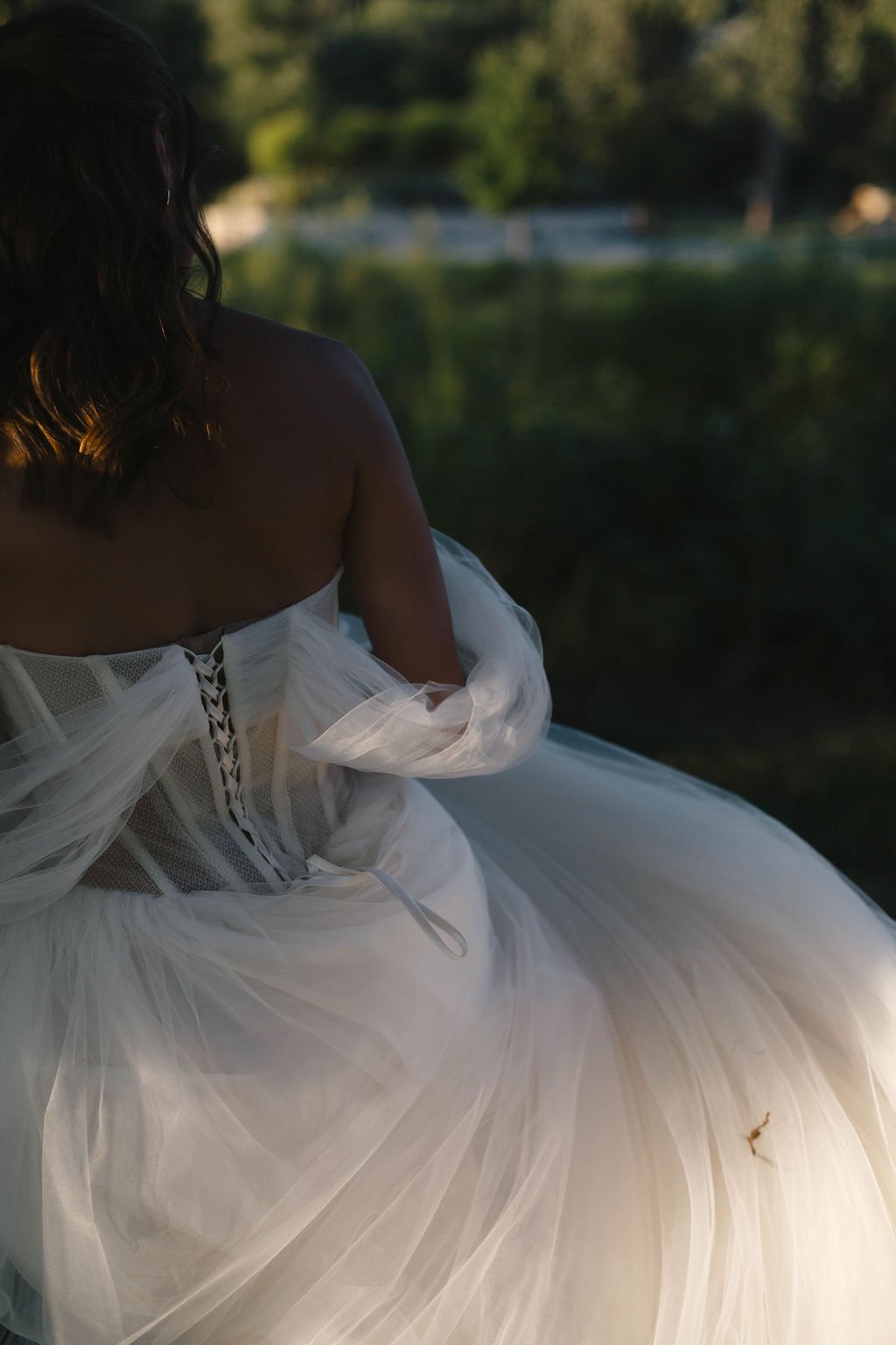 A woman in a strapless white wedding dress sitting outdoors near a body of water, with trees in the background, during what appears to be sunset.