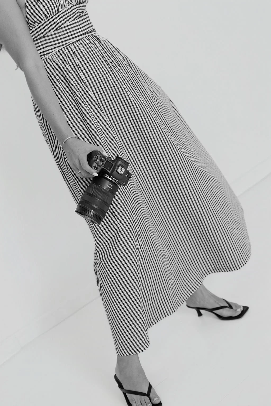 A woman in a striped dress and high heels holding a camera, standing against a plain white background.