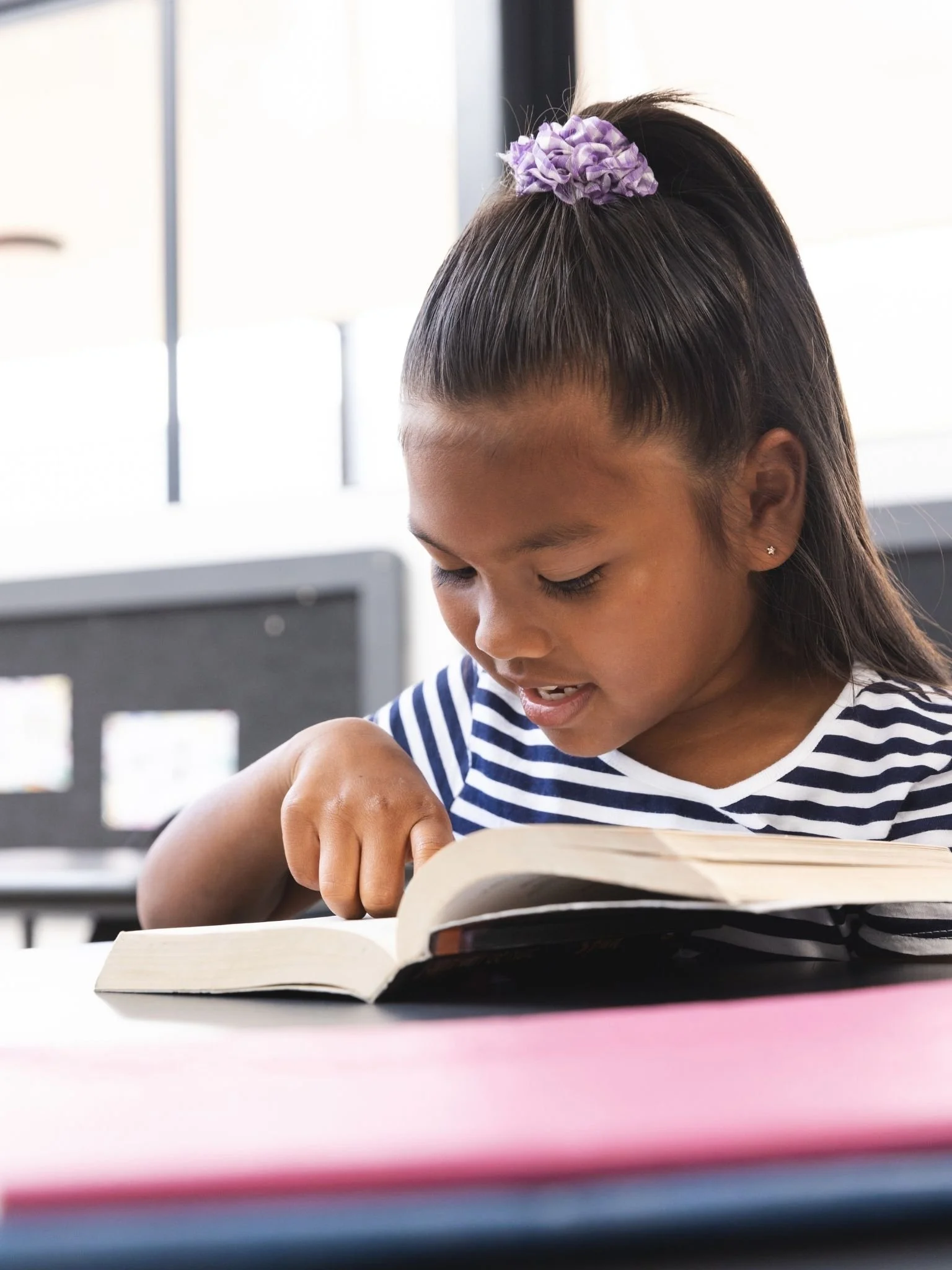 A student follows the text while reading a book at a classroom table.