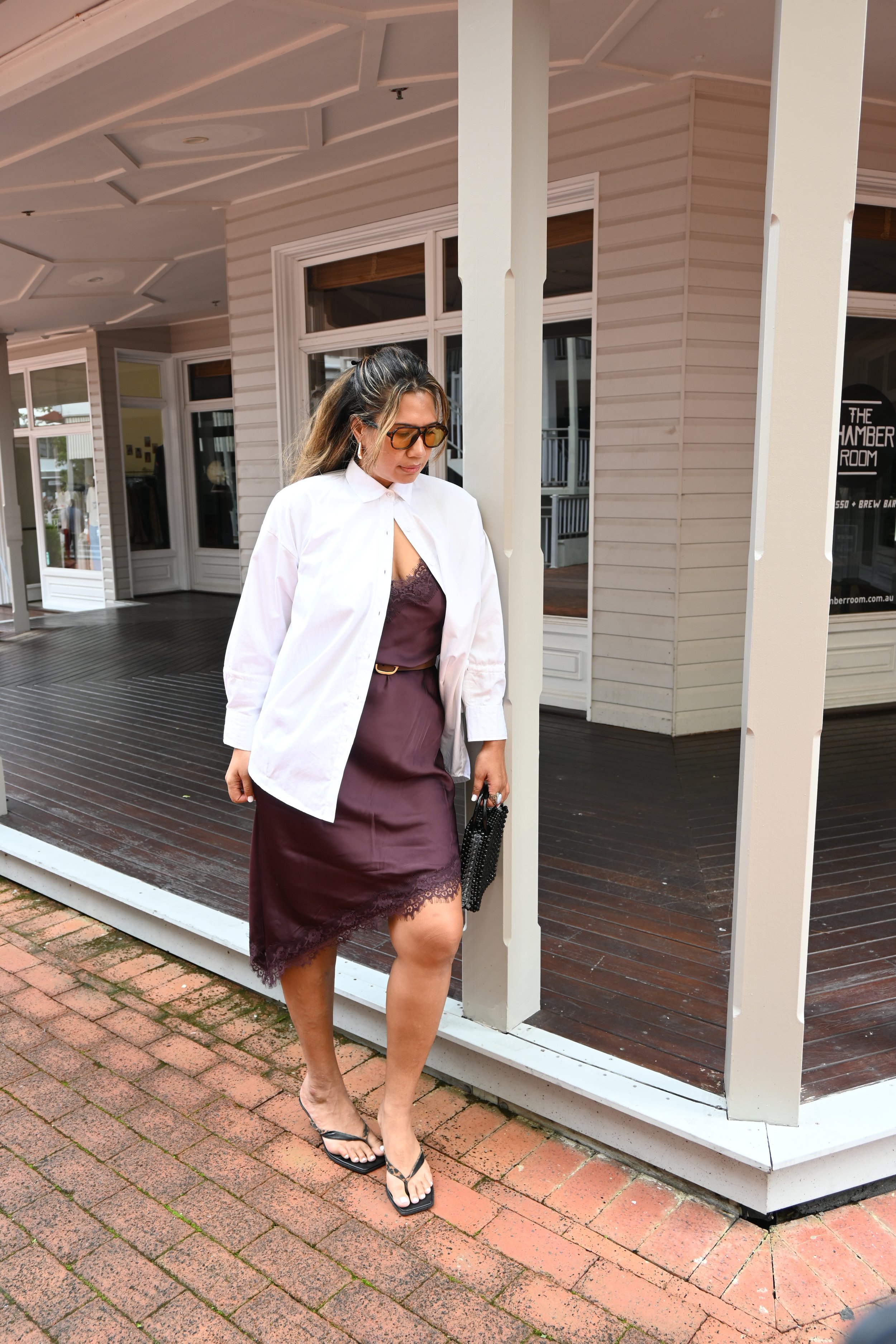 A woman in a white shirt, purple dress, and flip-flops walking on brick pavement outside a building with wooden siding and large windows.
