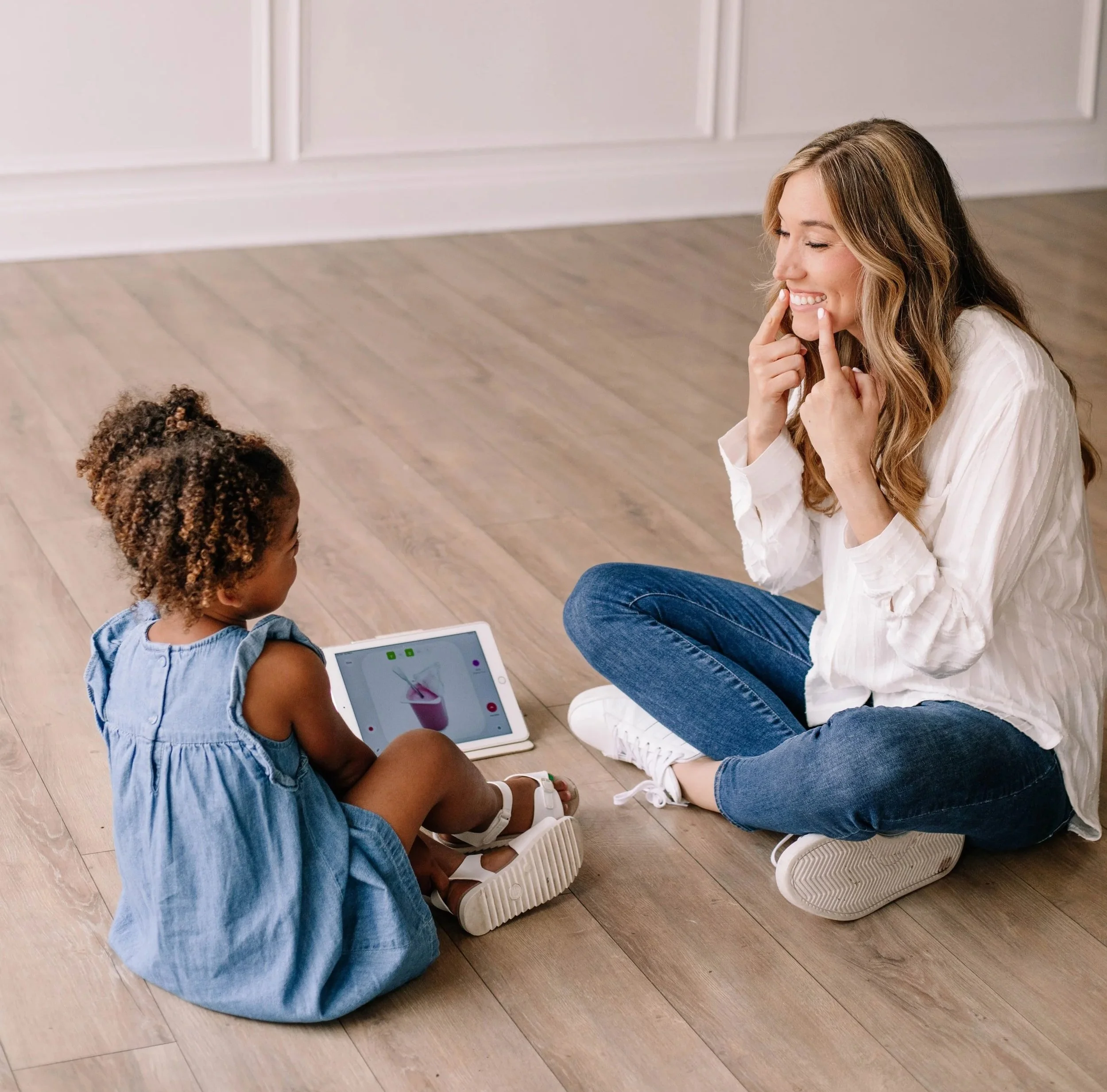 A myofunctional therapist is sitting on the floor with a young girl, smiling and touching her face, while working on tongue- and lip-tie exercises during myofunctional therapy in Dallas, TX.