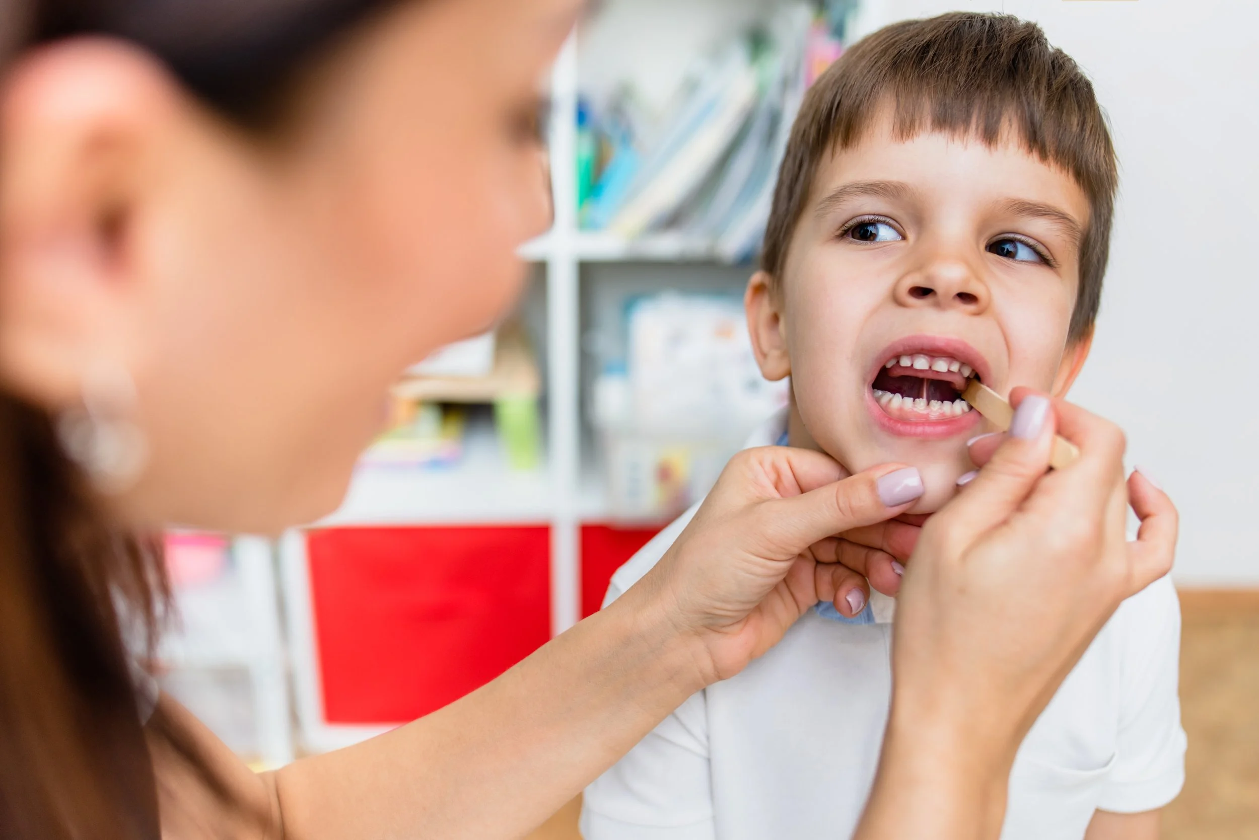 A young boy with brown hair participating in myofunctional therapy in Richardson, TX. The therapist is using a wooden tongue depressor for mouth and tongue exercises.