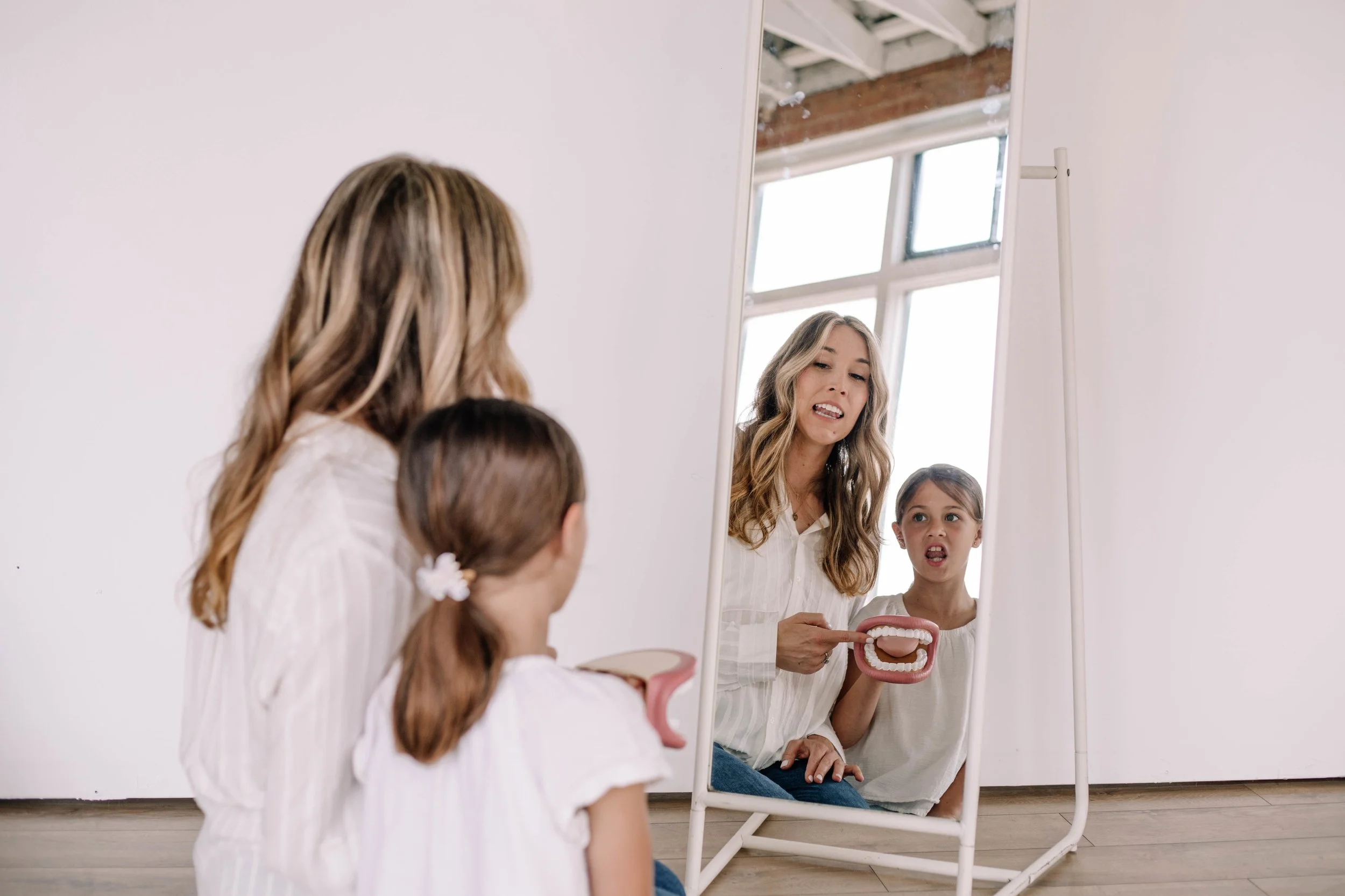 An orofacial myologist and a young girl sitting in front of a mirror, smiling and holding a dental model of teeth, working on tongue placement for correct speech sounds during myofunctional therapy in Richardson, TX.