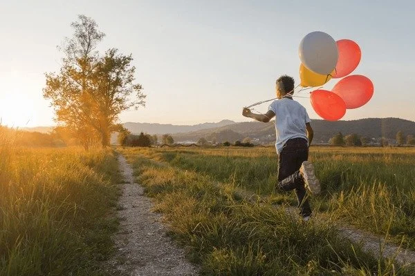 Child running on a dirt path holding colorful balloons in a grassy field at sunset.