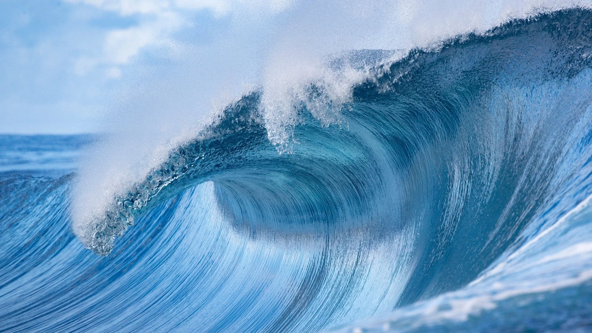 Photo of a large ocean wave with water spraying off the crest, set against a background of blue sky and distant horizon.
