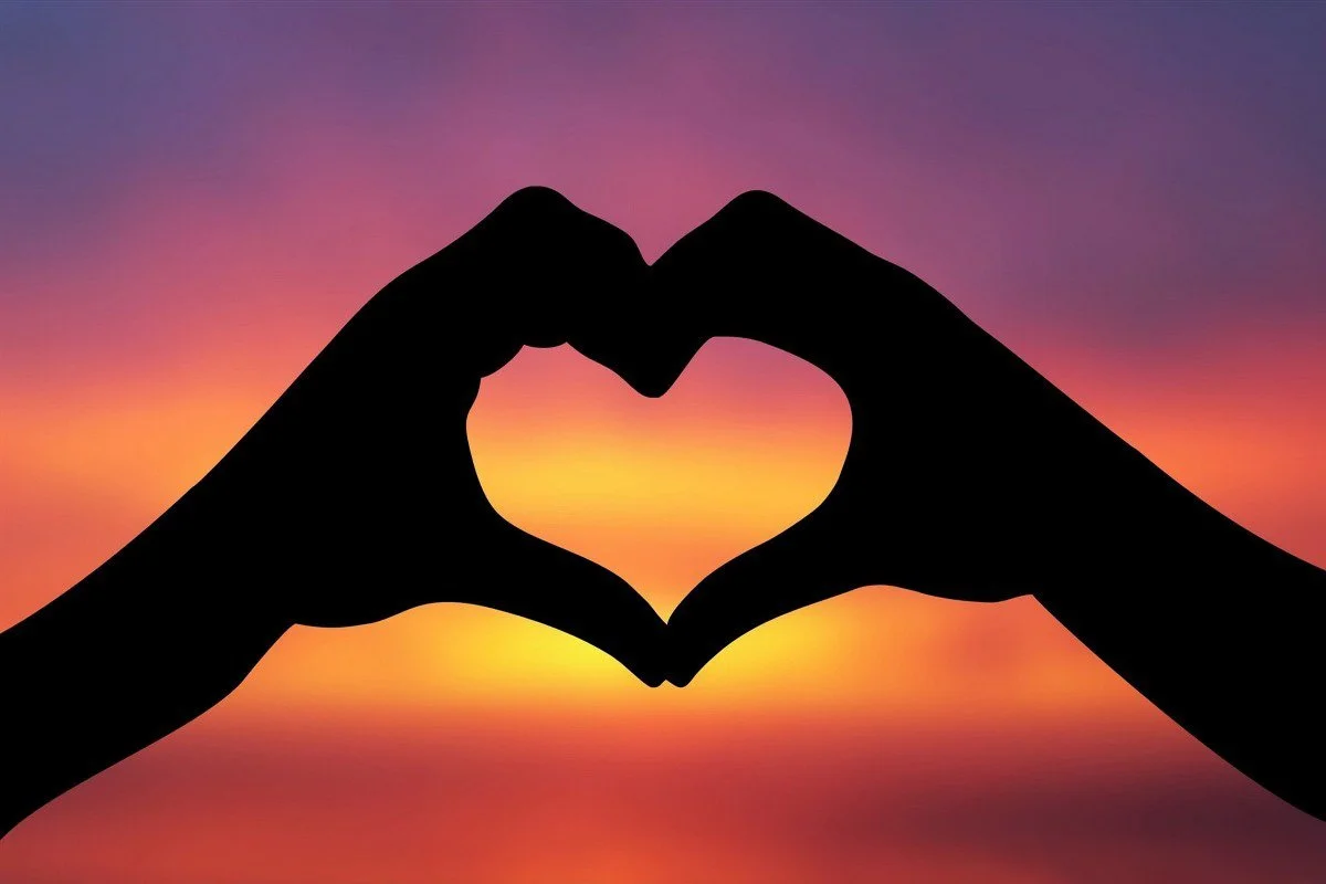 Silhouettes of two hands forming a heart shape against a colorful sunset sky with shades of purple, pink, orange, and yellow.