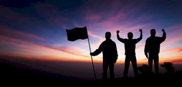 Silhouettes of three people celebrating on a hilltop during sunset, with one holding a flag and the other two raising their fists.