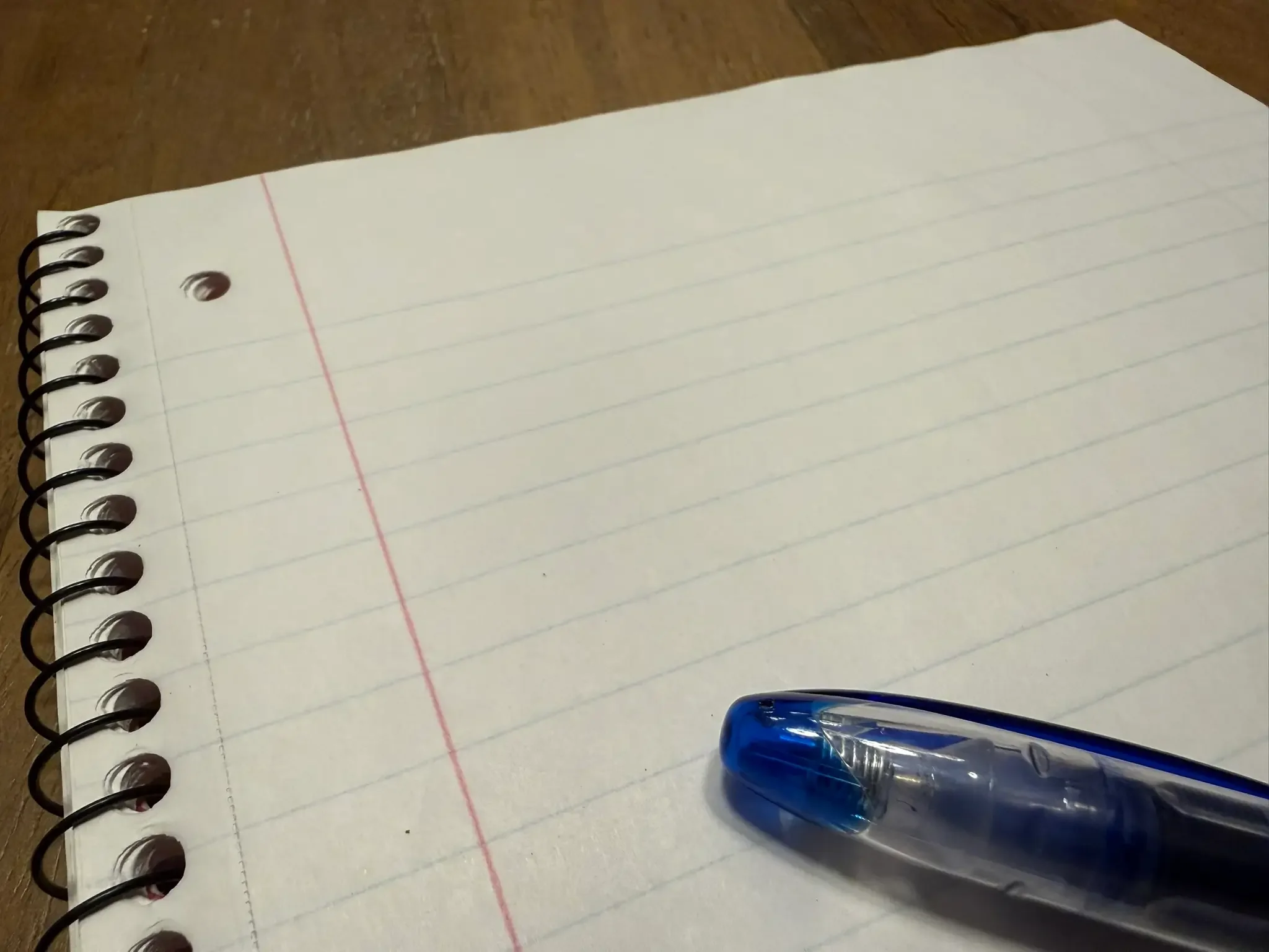Close-up of an empty lined notebook page with a blue pen placed on the bottom right corner, on a wooden surface.