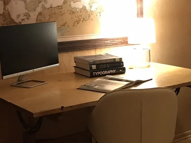 A neatly arranged wood drafting desk with a computer monitor, a stack of books design books, a lamp providing warm, cozy light.