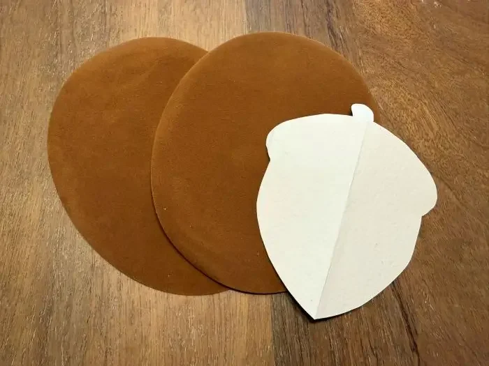 Two circular brown suede elbow pads and a white cardstock cutout of an acorn.