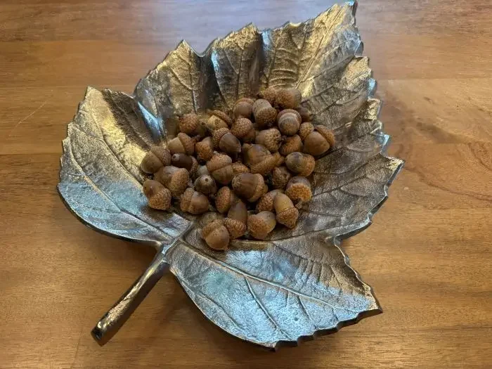 Decorative silver maple leaf-shaped dish filled with acorns, placed on a wooden surface.