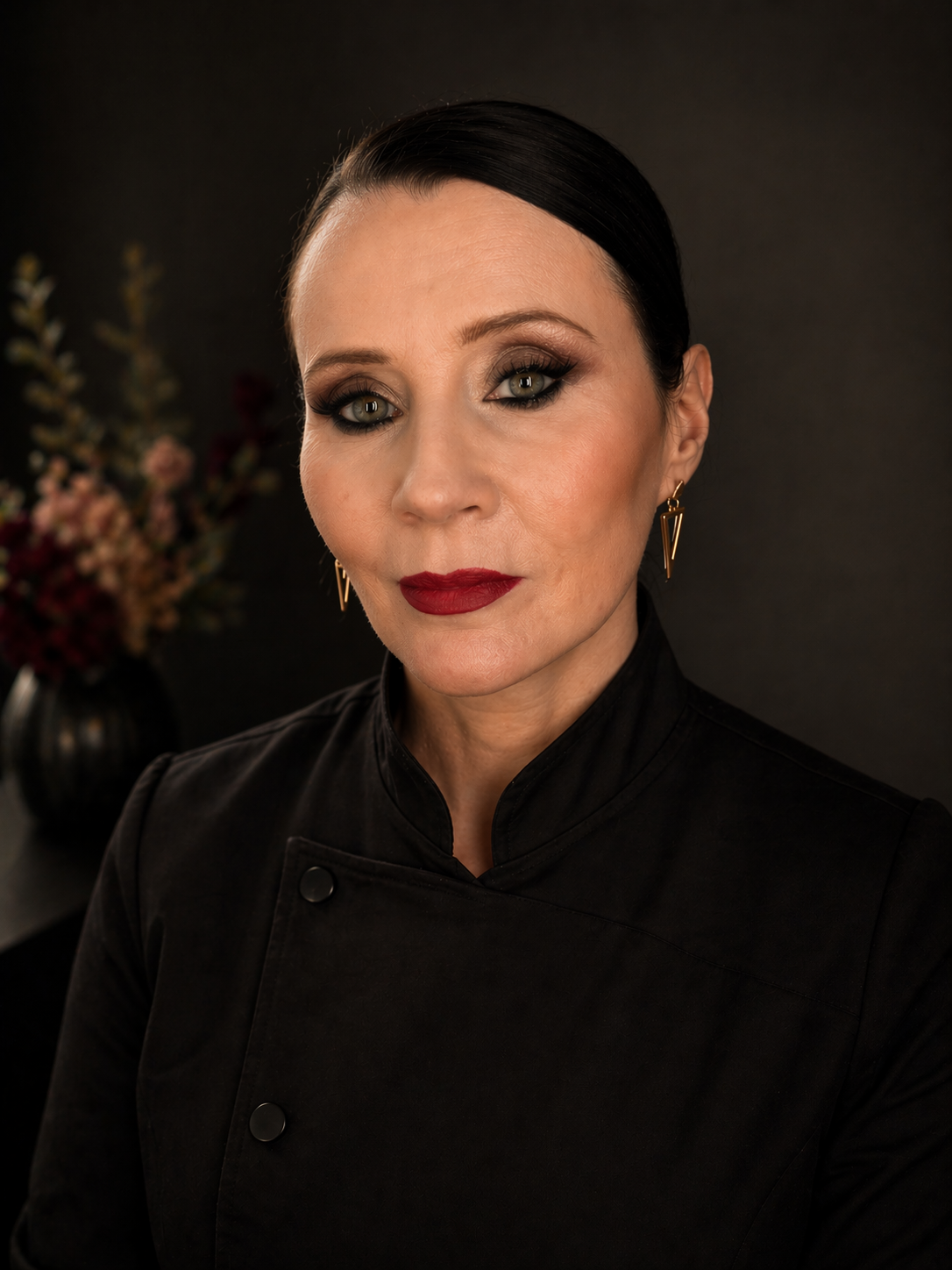 A woman with dark hair and makeup wearing a black chef's coat, posing in front of a dark background with a vase of flowers.