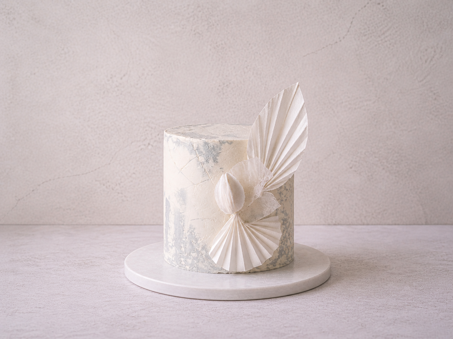 White marble cake on a round marble base with white decorative paper art on the side, set against a light textured wall.