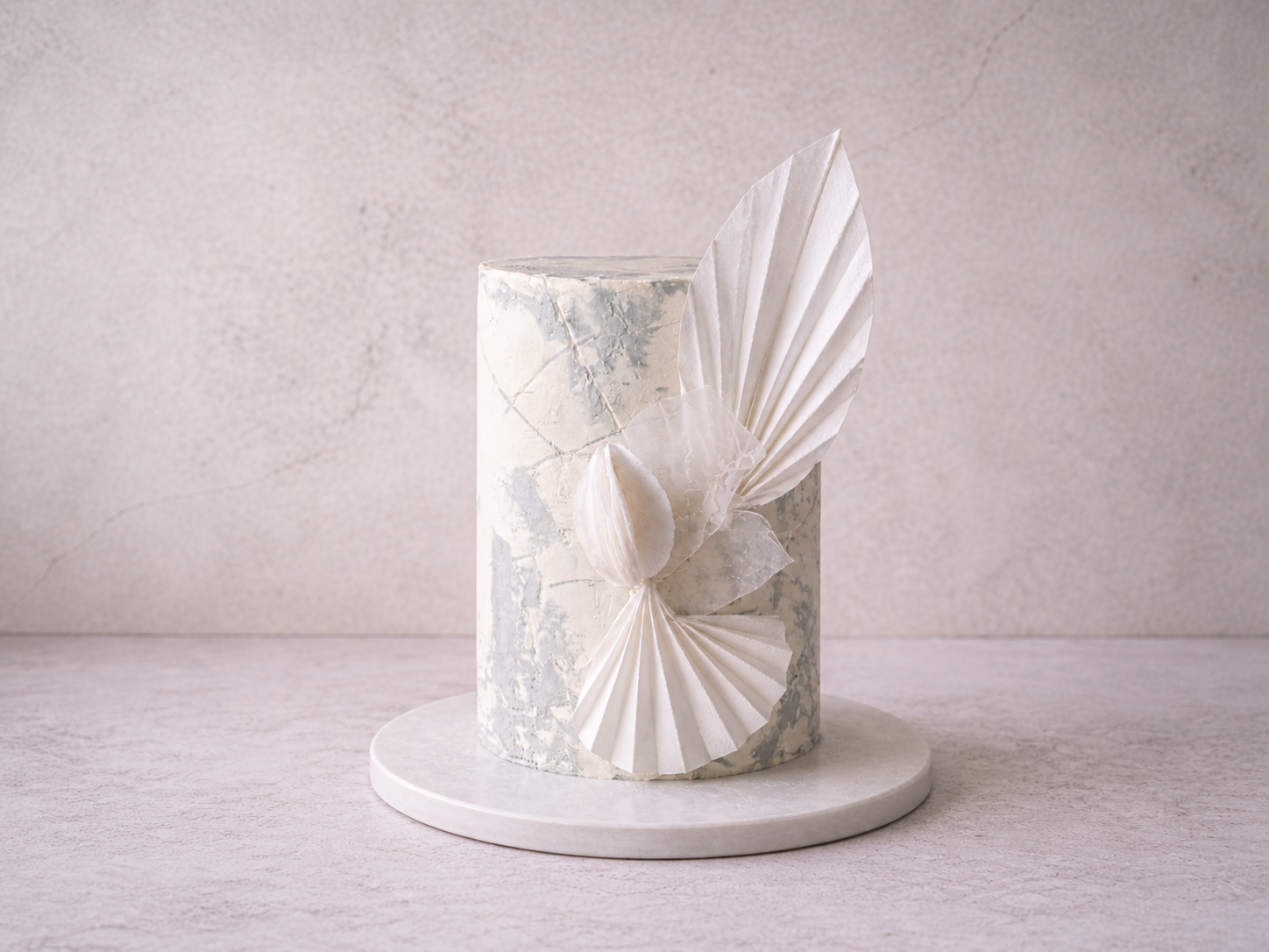 A tall cylindrical cake with a marbled white and gray design, decorated with white paper fan and shell embellishments, on a round white base against a neutral background.