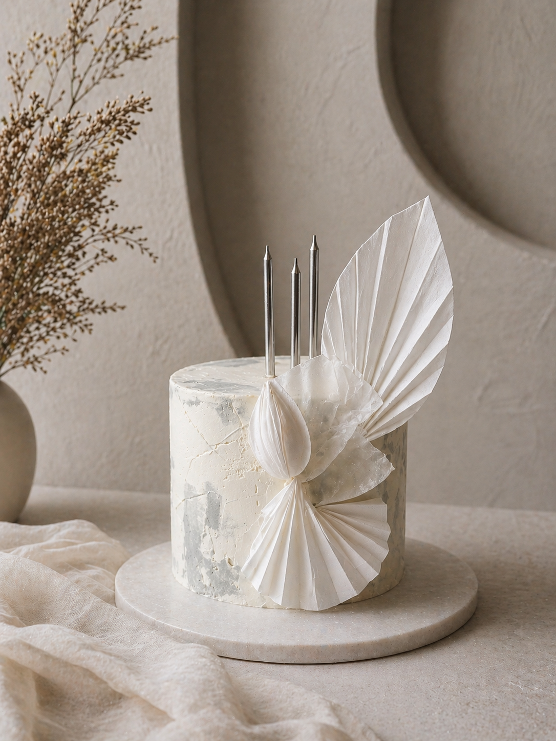 White marble cake with decorative white paper fans and silver candles on a round base, with beige background and dried plant in a vase.
