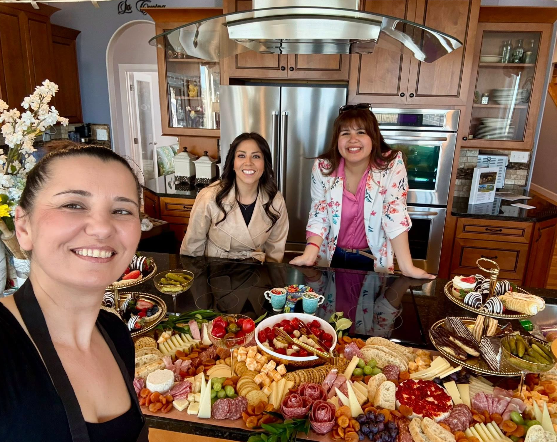 Four women smiling in a kitchen surrounded by an assortment of appetizers and desserts on a table.