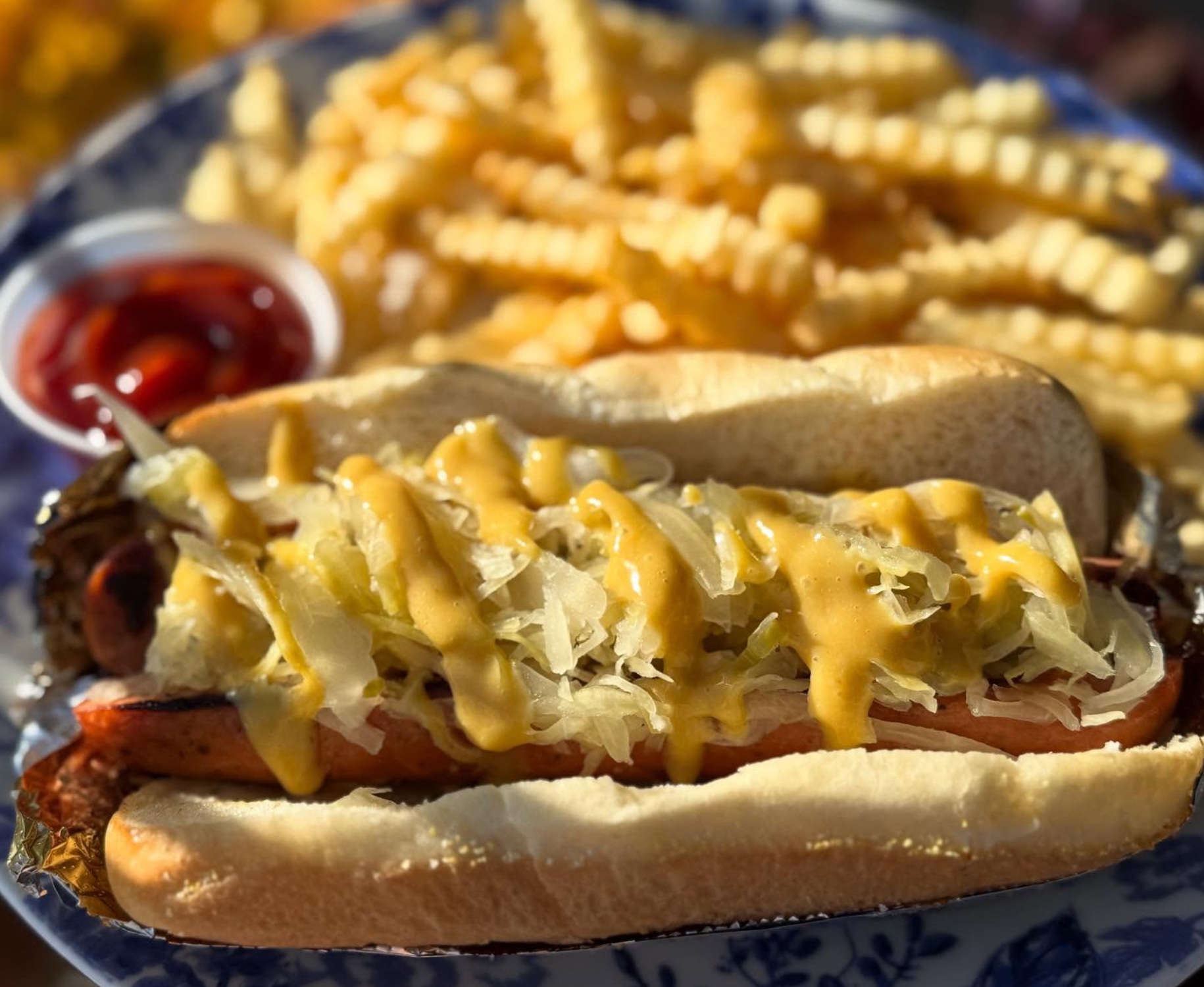 Close-up of a hot dog in a bun topped with shredded cheese and mustard, with a side of ketchup and a waffle fry in the background.
