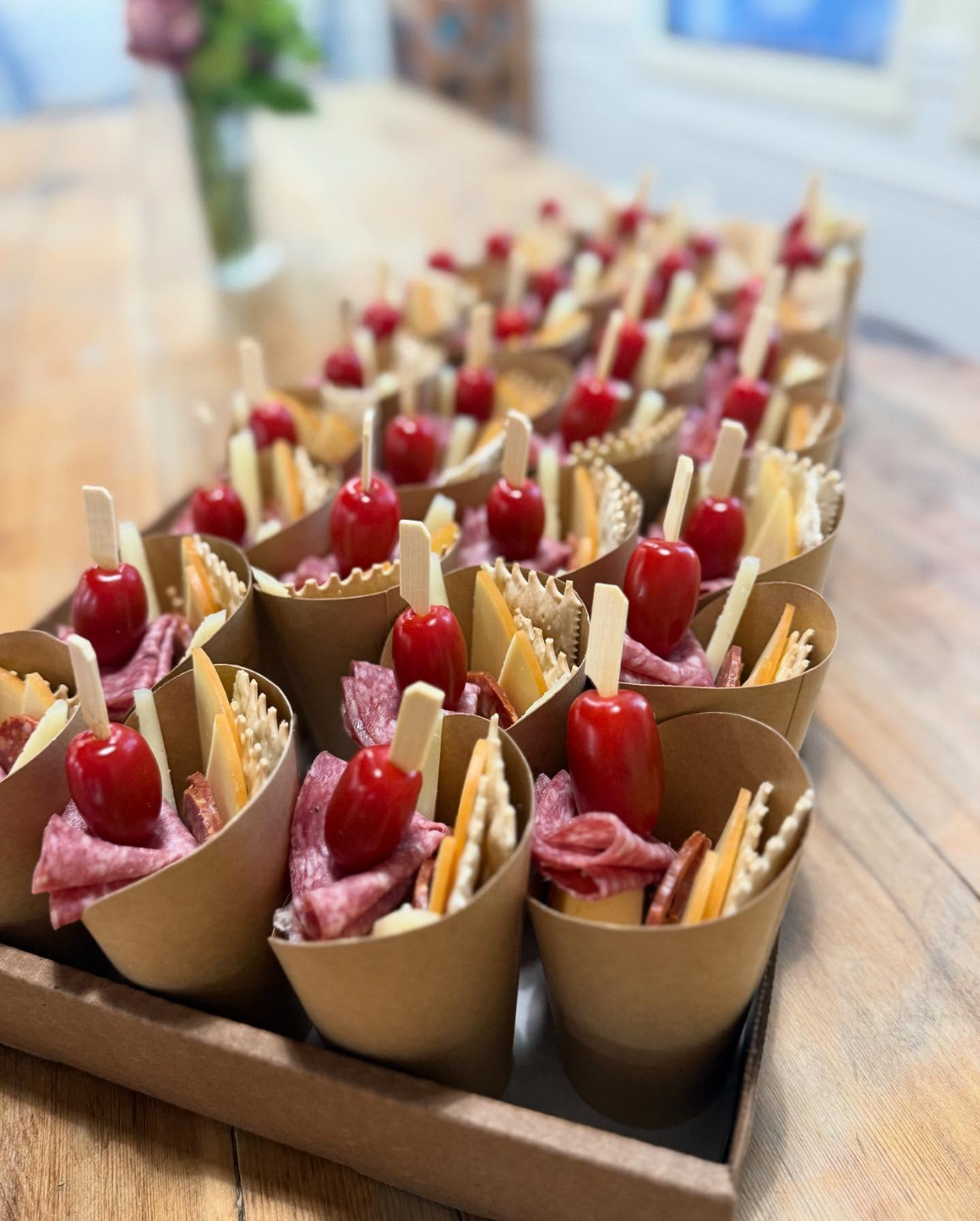 Tray of party appetizers with cheese, sliced meats, cherry tomatoes on sticks, and crackers served in cone-shaped paper holders.