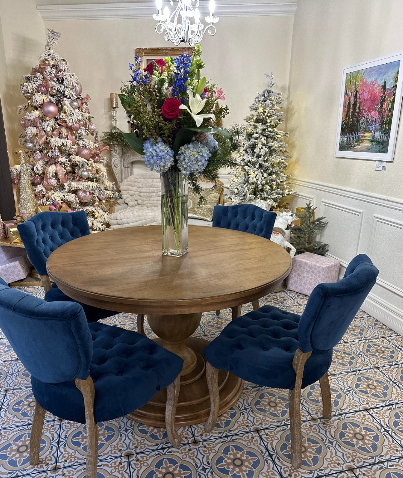 A round wooden dining table with five blue upholstered chairs, a tall glass vase with a large floral arrangement, Christmas trees decorated with ornaments and snow effects, a chandelier, a colorful framed painting, and wrapped gifts.