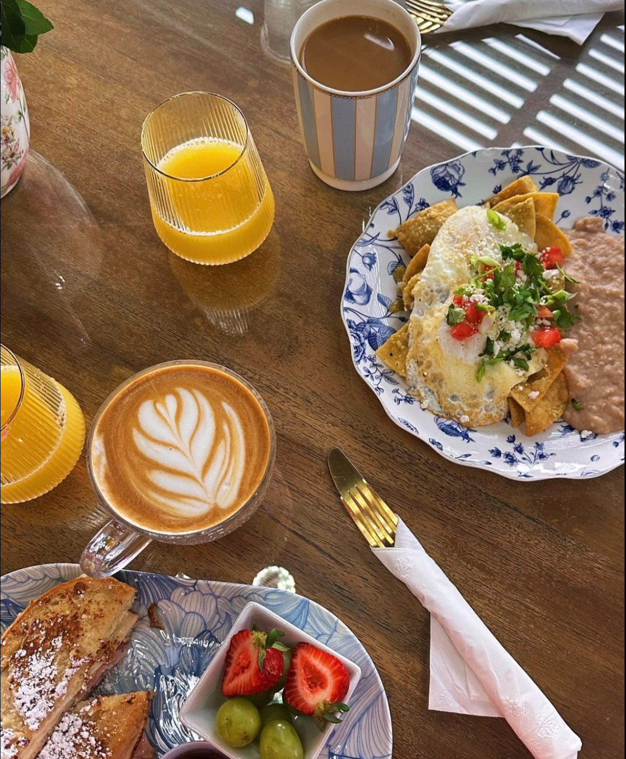 A breakfast scene with a plate of Mexican food, a cup of coffee with latte art, a glass of orange juice, a mug of coffee with milk, and a plate of strawberries and grapes on a wooden table.