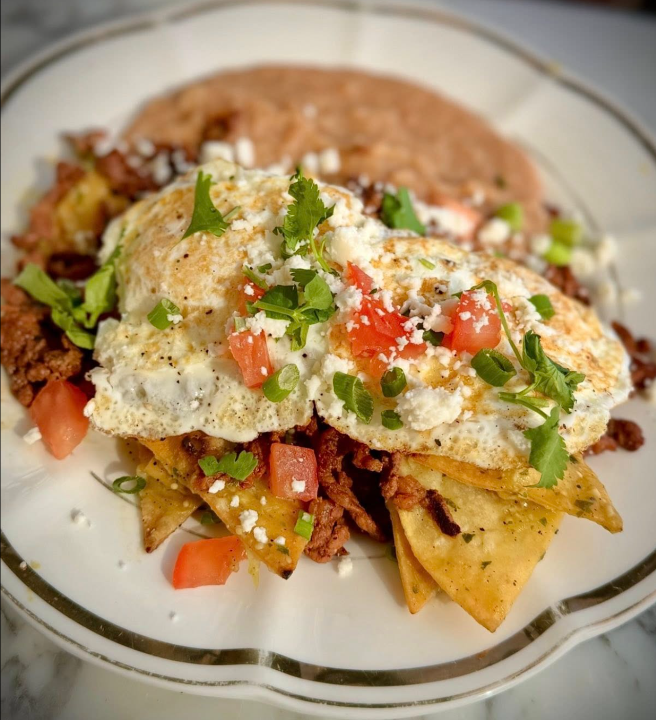 Plate of Mexican nachos topped with fried eggs, chopped tomatoes, green onions, cilantro, and crumbled cheese.