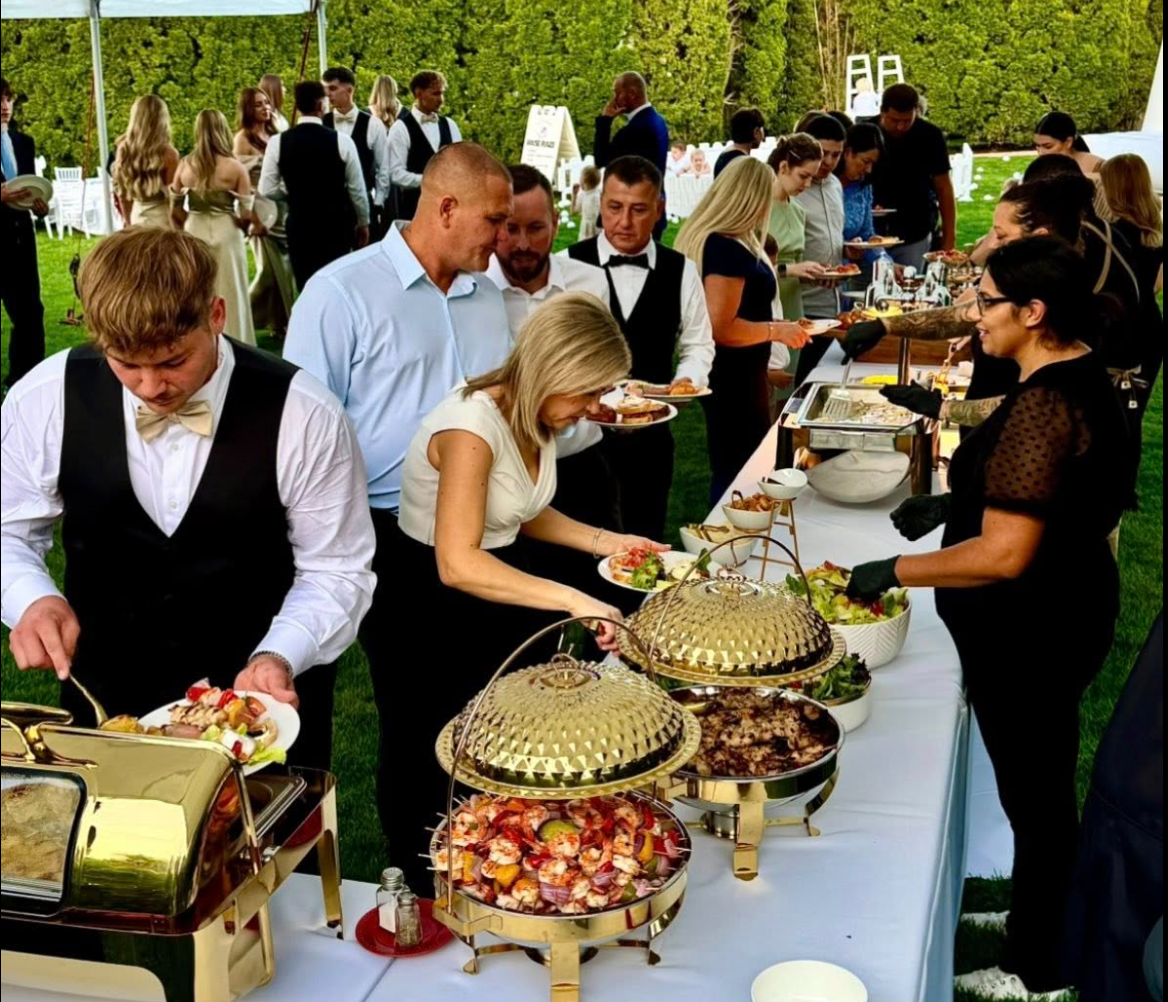 Guests at an outdoor buffet table at a formal event, serving themselves food under a white canopy with trees in the background.