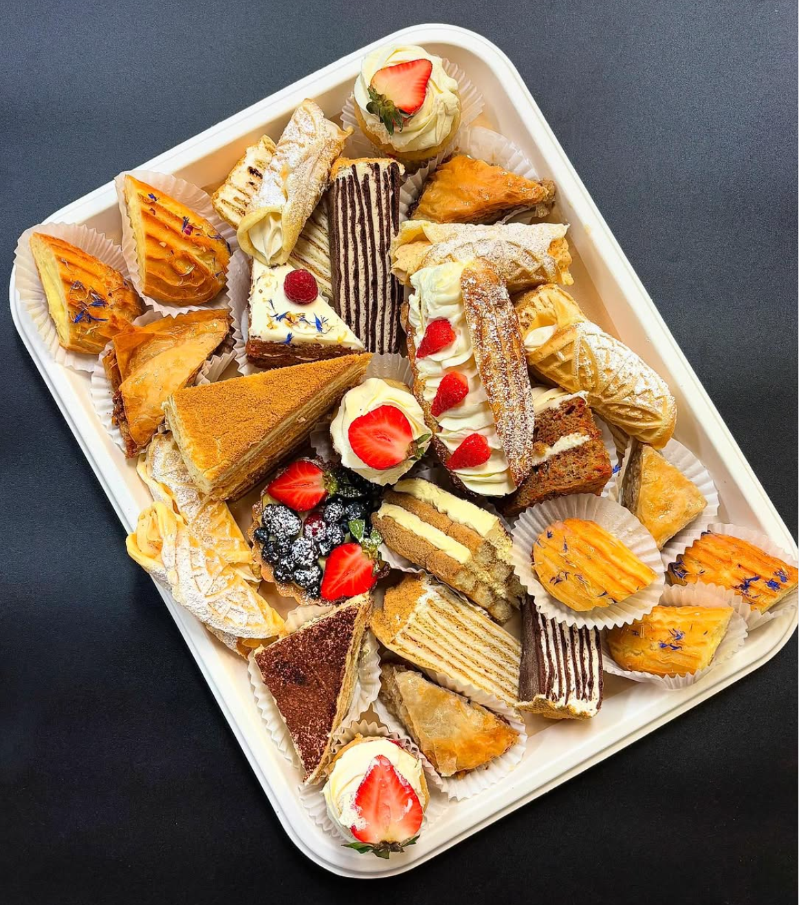Assorted slices of various cakes and pastries on a white tray, decorated with strawberries, blueberries, raspberries, and edible flowers.