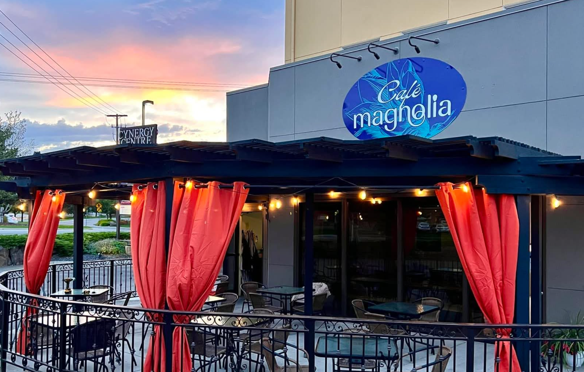 Exterior view of Cafe Magnolia restaurant with red curtains, outdoor seating, and a blue sign reading 'Café Magnolia' at sunset.