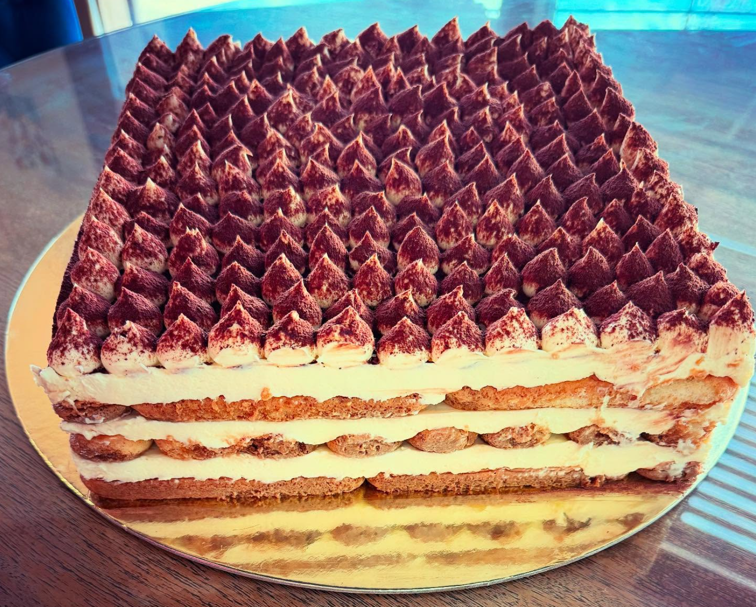 A layered tiramisu cake with a top decorated with piped mascarpone and cocoa powder, placed on a gold-colored base