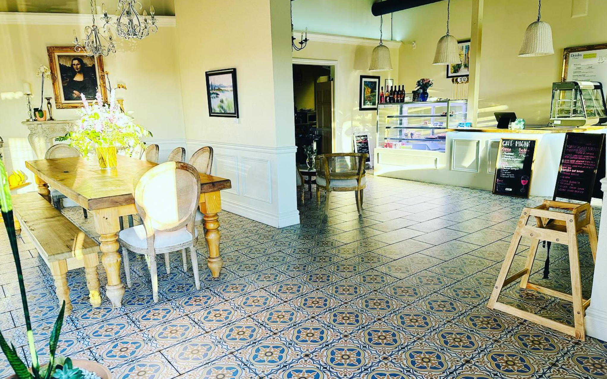 Interior of a bright cafe with a wooden dining table and chairs, decorative tiled floor, framed artwork on yellow walls, chandelier, and a counter area with bottles and menu boards.