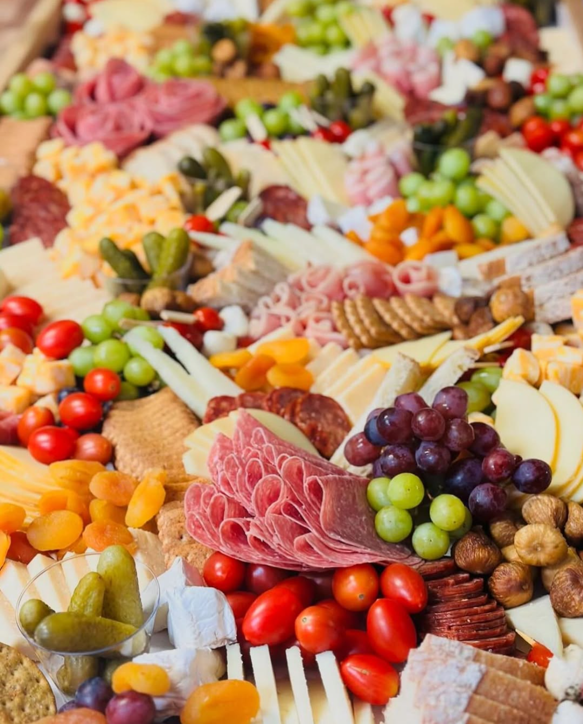 A large platter of assorted cheeses, grapes, cherry tomatoes, cured meats, pickles, crackers, and dried fruits.