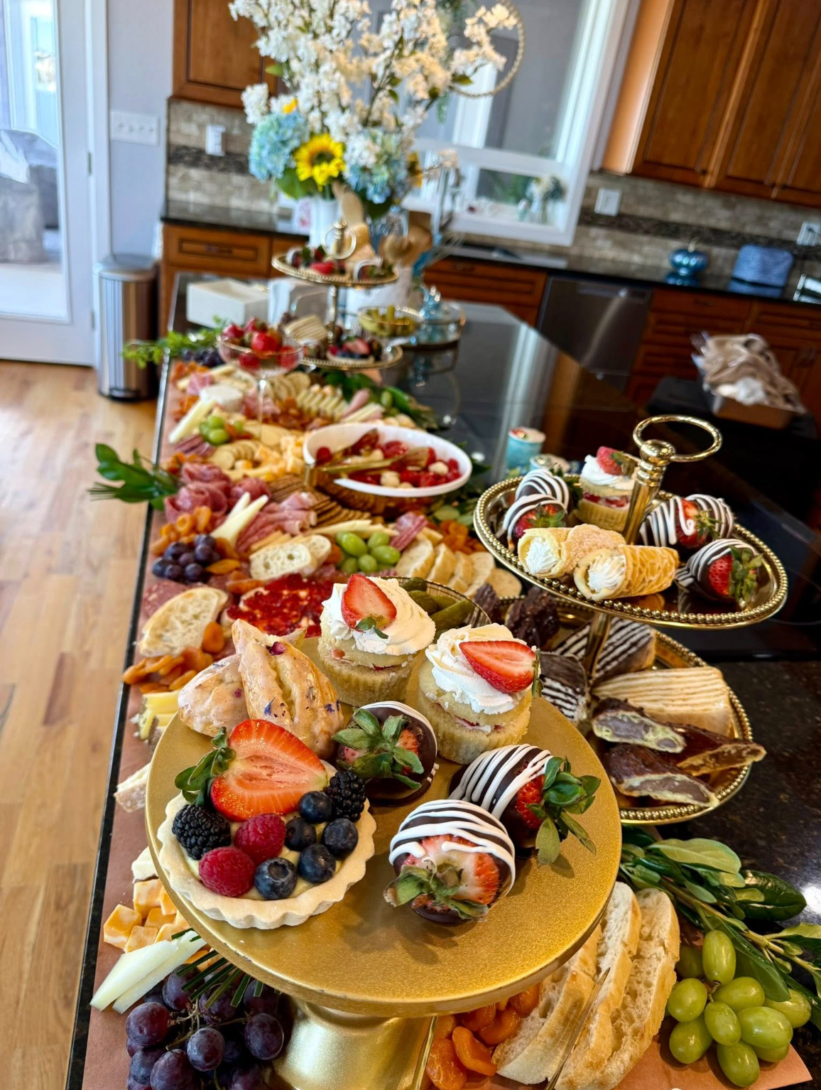 Assorted desserts and snacks on a table, including cupcakes with strawberries, chocolate-covered strawberries, fruit tarts, and a variety of finger foods, set in a home kitchen with flowers and a window in the background.