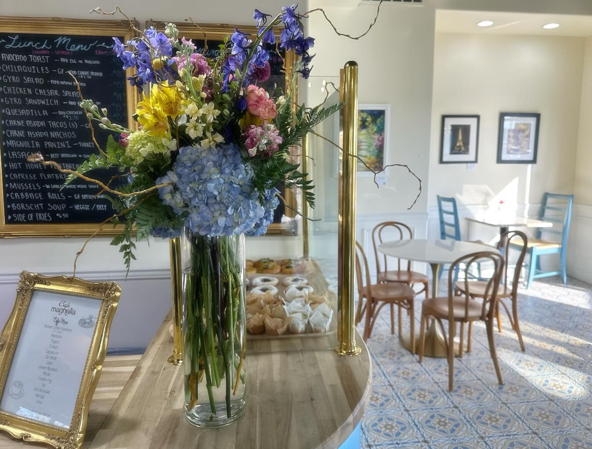 A tall glass vase with a large bouquet of colorful flowers, including blue hydrangeas, pink ranunculus, white, and purple flowers, on a wooden table in a bright, decorated room with chairs, paintings on the wall, and a chalkboard menu in the background.